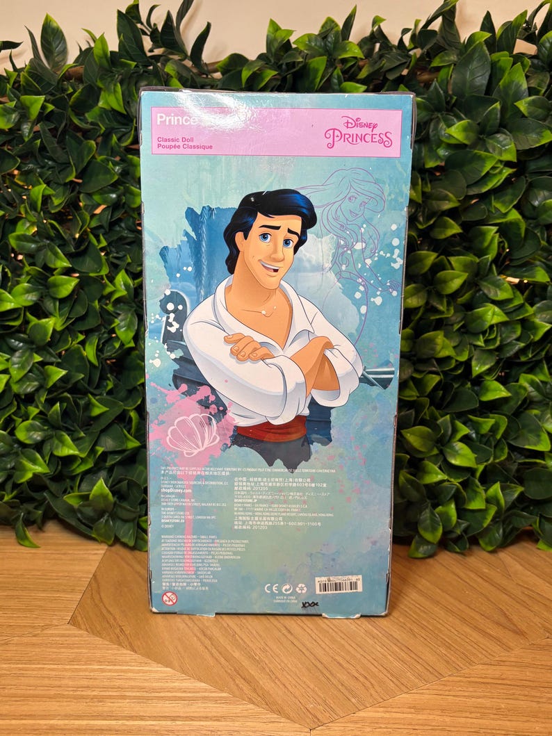 May include: A Disney Princess Prince doll box featuring a cartoon illustration of a man with black hair, wearing a white shirt and red sash. The box has the words "Prince" and "Classic Doll" printed on it.