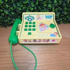 Peut inclure: Un téléphone jouet jaune Cabbage Patch Kids avec un combiné et un cordon verts. L'appareil dispose d'un clavier à touches vertes, d'une illustration centrale du personnage Georgia Ann et de dessins d'enfants sur le bord avant du boîtier.