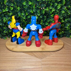 May include: Three superhero action figures: Wolverine in yellow and blue, Captain America in blue, white, and red, and Spider-Man in red and blue. They are arranged on a wooden oval tray against a green leafy background.