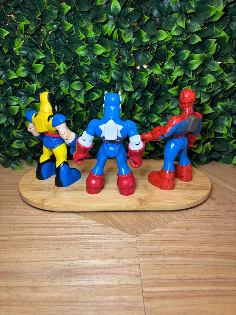 May include: Three superhero action figures: Wolverine in yellow and blue, Captain America in blue, white, and red, and Spider-Man in red and blue. They are arranged on a wooden oval tray against a green leafy background.
