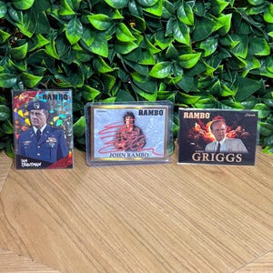 May include: Three Rambo trading cards displayed side-by-side. The left card features Sam Trautman in a military uniform. The centre card shows John Rambo with a red signature overlay. The right card depicts Robert Griggs against an explosion.