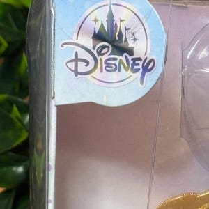 May include: A clear plastic package with a Disney logo sticker. The sticker features a castle silhouette, stars, and the word "Disney" in a stylized font. The package contains a doll or toy.
