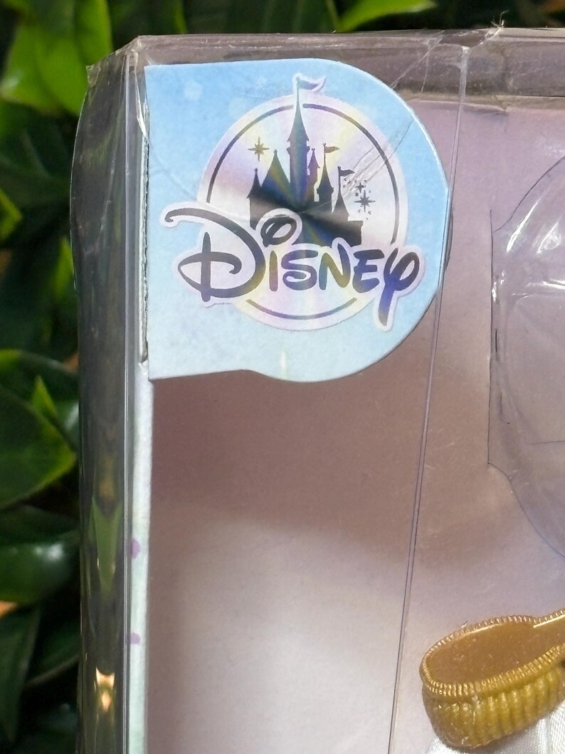 May include: A clear plastic package with a Disney logo sticker. The sticker features a castle silhouette, stars, and the word "Disney" in a stylized font. The package contains a doll or toy.