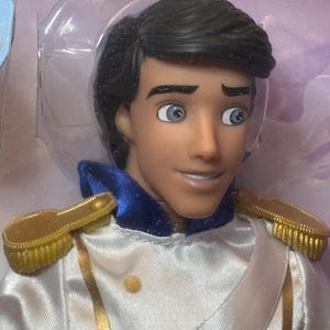 May include: A doll of a prince with black hair, blue eyes, and a tan complexion. He wears a white satin jacket with gold trim and epaulets, and a blue collar. The doll is in a clear plastic package.