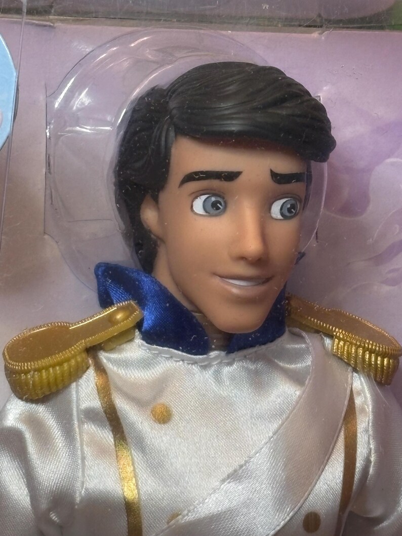 May include: A doll of a prince with black hair, blue eyes, and a tan complexion. He wears a white satin jacket with gold trim and epaulets, and a blue collar. The doll is in a clear plastic package.
