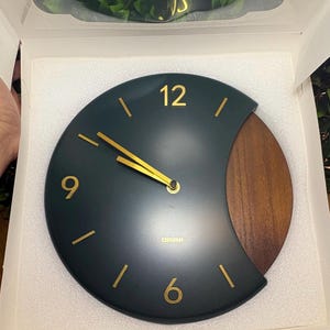 Peut inclure: Une horloge murale ronde avec un cadran vert foncé, des aiguilles et des chiffres dorés aux positions 6, 9 et 12. Le côté droit du cadran présente un panneau en bois en forme de croissant. La marque ORIINI est imprimée en or sous les aiguilles.