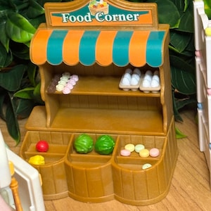 May include: A miniature brown plastic food stand with a teal and orange striped awning and a sign reading Food Corner. The stand features shelves stocked with small plastic eggs, colorful round treats, and green produce items.