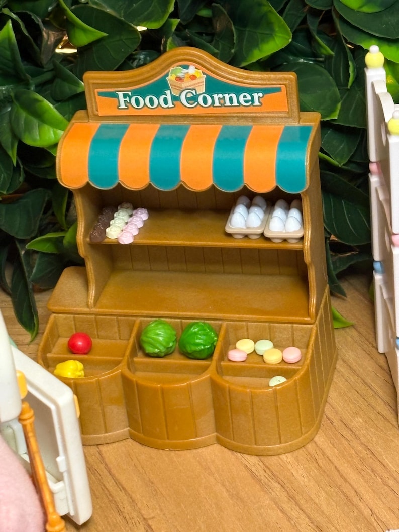 May include: A miniature brown plastic food stand with a teal and orange striped awning and a sign reading Food Corner. The stand features shelves stocked with small plastic eggs, colorful round treats, and green produce items.