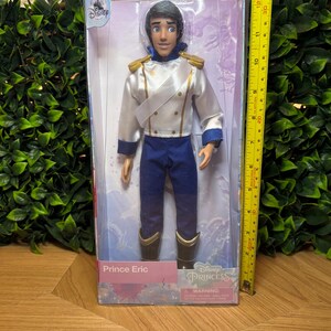May include: A Prince Eric doll in a clear plastic box. The doll is wearing a white jacket with gold accents, blue pants, and black boots. The box is labeled "Disney Princess" and "Prince Eric."