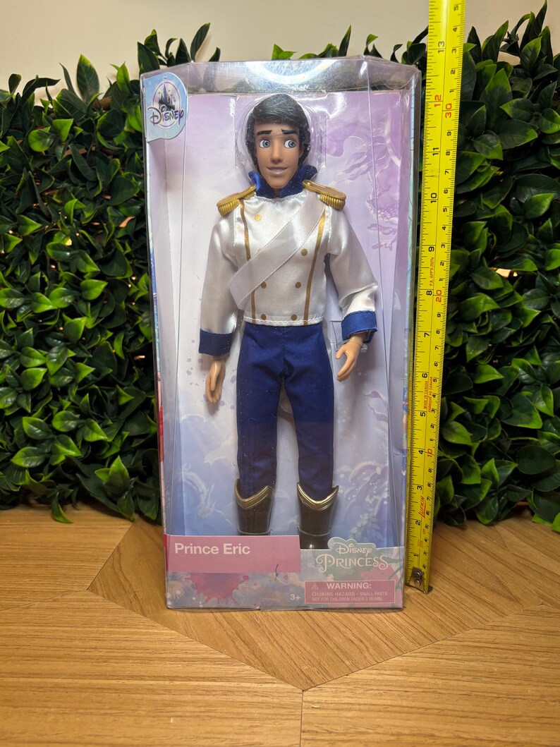 May include: A Prince Eric doll in a clear plastic box. The doll is wearing a white jacket with gold accents, blue pants, and black boots. The box is labeled "Disney Princess" and "Prince Eric."