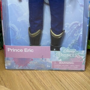 May include: A Prince Eric doll in a box. The doll is wearing blue pants and black boots with gold trim. The box has the words "Prince Eric" and "Disney Princess" printed on it. A warning label is also visible.