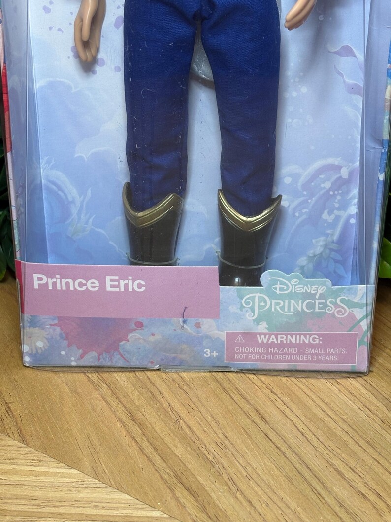 May include: A Prince Eric doll in a box. The doll is wearing blue pants and black boots with gold trim. The box has the words "Prince Eric" and "Disney Princess" printed on it. A warning label is also visible.