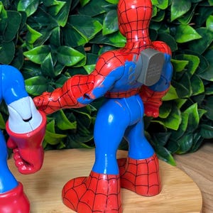 May include: A red, blue, and black Spider-Man action figure. The figure has a red head and torso with a black web pattern, blue arms and legs, and red webbed feet. The figure is standing on a wooden surface.