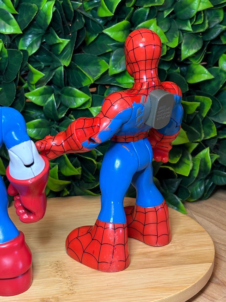 May include: A red, blue, and black Spider-Man action figure. The figure has a red head and torso with a black web pattern, blue arms and legs, and red webbed feet. The figure is standing on a wooden surface.