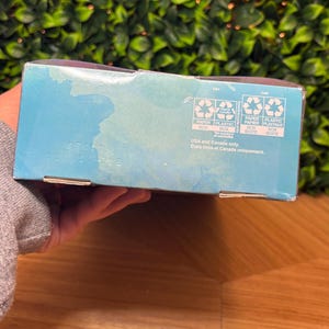 May include: A partially visible, light blue box with recycling symbols and text. The box has text in English and French, indicating it's for use in the USA and Canada. The background features green foliage and a wooden surface.