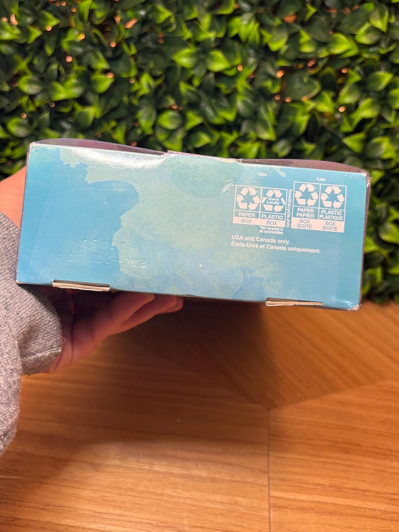May include: A partially visible, light blue box with recycling symbols and text. The box has text in English and French, indicating it's for use in the USA and Canada. The background features green foliage and a wooden surface.