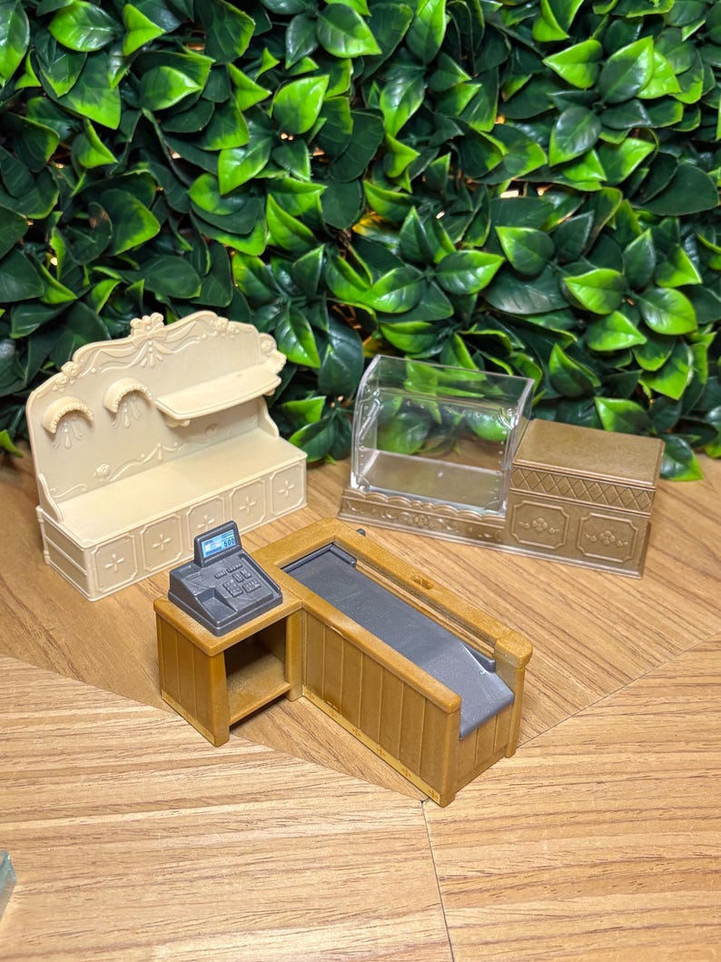 May include: A miniature retail set featuring a beige wall shelf unit with decorative molding, a clear plastic display case with a gold base, and a brown wooden checkout counter equipped with a small gray cash register and a conveyor belt.