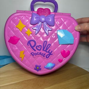 May include: A pink Polly Pocket heart-shaped carrying case with a quilted pattern. It features a purple handle, bow, and decorative accents including a diamond, stars, and the Polly Pocket logo. A blue strap is visible.