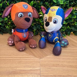 May include: Two plush dog toys, one orange and brown, the other blue and white. The orange dog has a helmet and uniform with an anchor emblem. The blue dog has a helmet and uniform with a star emblem. Both toys are on a wooden surface.