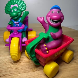 May include: Two colorful toy vehicles featuring Barney and Baby Bop characters. Barney is in a red wagon, and Baby Bop is on a purple tricycle. Both toys have yellow wheels and are made of plastic.