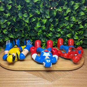 May include: Four superhero action figures: Wolverine in yellow and blue, Captain America in blue, red, and white, and Spider-Man in red and blue. They are lying on a wooden tray against a backdrop of green leaves.