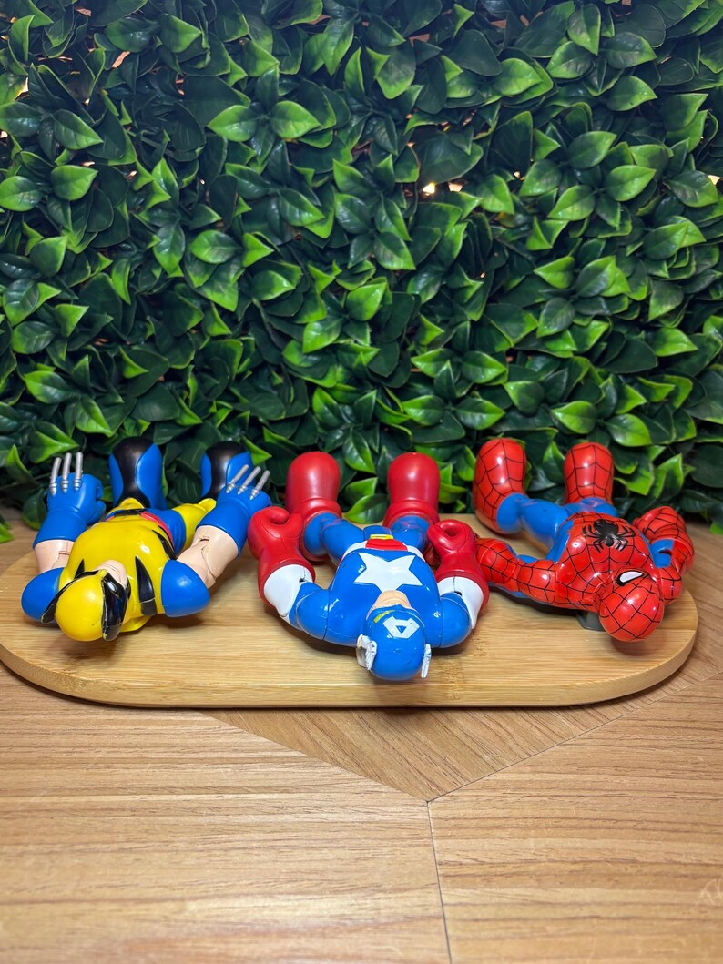 May include: Four superhero action figures: Wolverine in yellow and blue, Captain America in blue, red, and white, and Spider-Man in red and blue. They are lying on a wooden tray against a backdrop of green leaves.