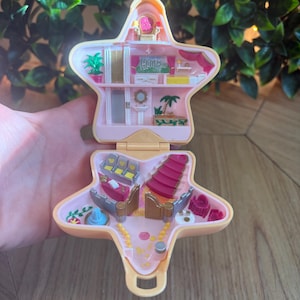 May include: A star-shaped plastic playset that opens to reveal a miniature interior with two levels. The top level features a room with a Hollywood sign, a plant, and a bed. The bottom level includes theater-style seating, a staircase, and small furniture pieces.