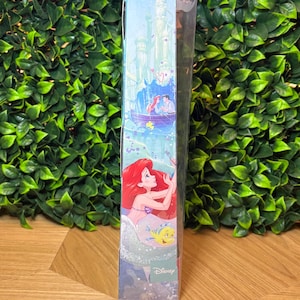 May include: A Disney-themed box featuring illustrations of Ariel, Prince Eric, and Flounder. The box has a light blue background with a castle and boat scene. The Disney logo is visible.