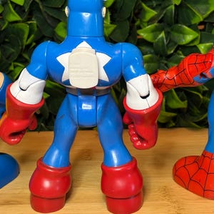 May include: A blue Captain America action figure with a white star on the chest, red gloves and boots. The figure is part of a set of superhero toys. The background is a wooden surface with green foliage.