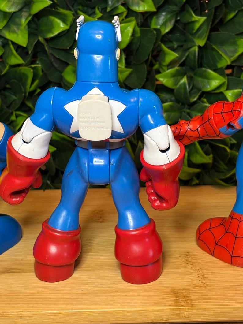 May include: A blue Captain America action figure with a white star on the chest, red gloves and boots. The figure is part of a set of superhero toys. The background is a wooden surface with green foliage.