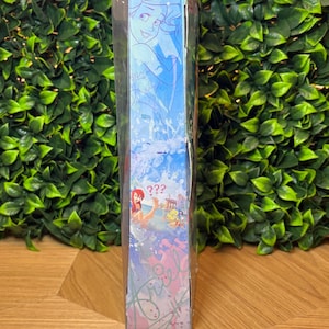 May include: A blue box featuring illustrations of Ariel from The Little Mermaid. The box has a light blue background with images of Ariel and other characters. The text "???" is visible on the box.