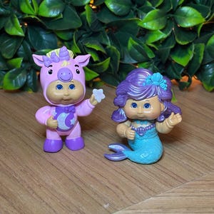 May include: Two small, colourful figurines. One is a pink unicorn-themed doll holding a star, the other is a mermaid with a blue tail and purple hair. Both dolls have large blue eyes and are on a wooden surface.