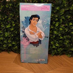 May include: A Disney Princess Prince Eric classic doll box. The box features an illustration of Prince Eric in a white shirt and a red sash. The box has the Disney Princess logo and the text "Prince Eric" in pink.