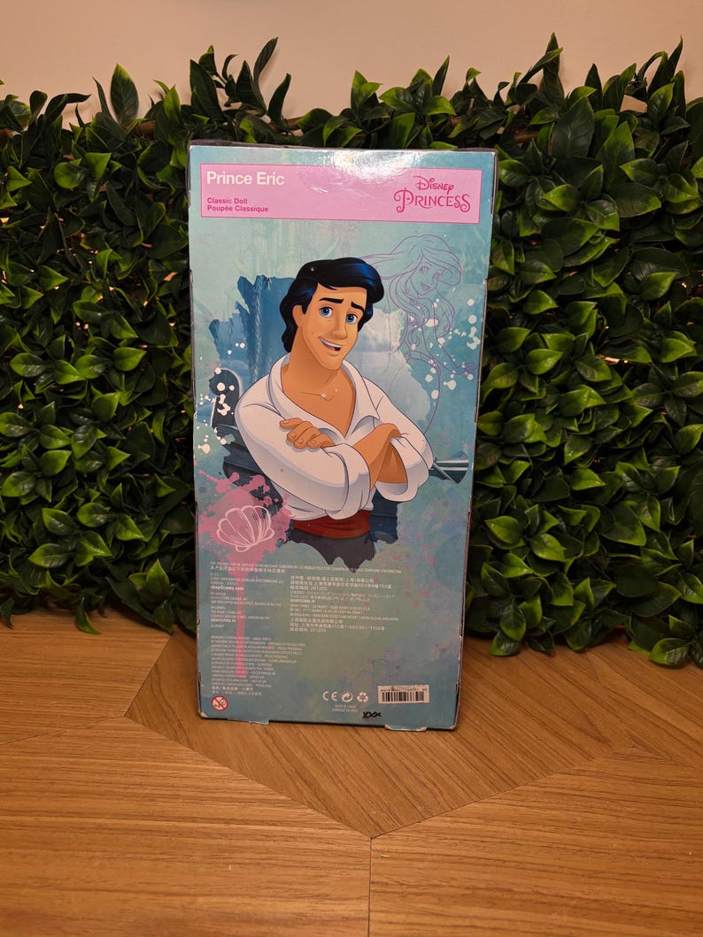 May include: A Disney Princess Prince Eric classic doll box. The box features an illustration of Prince Eric in a white shirt and a red sash. The box has the Disney Princess logo and the text "Prince Eric" in pink.