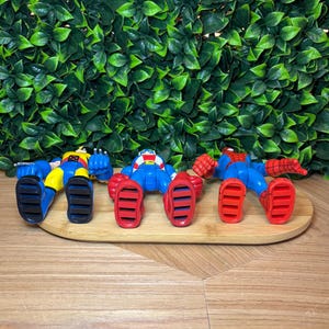 May include: Three superhero action figures in blue, red, and yellow, lying on a wooden tray. The figures have detailed features and are posed in a relaxed position. The background features a wall of green leaves.