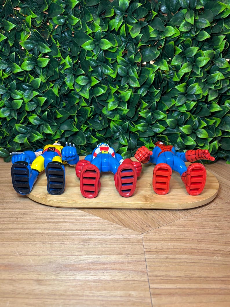May include: Three superhero action figures in blue, red, and yellow, lying on a wooden tray. The figures have detailed features and are posed in a relaxed position. The background features a wall of green leaves.