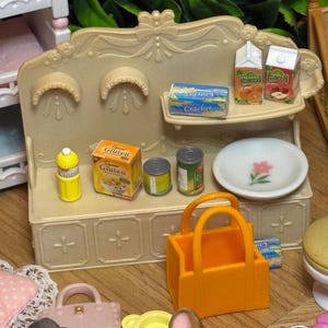 May include: A miniature beige kitchen hutch with a shelf holding two juice cartons and a cracker box. The counter features a yellow bottle, a gluten-free cereal box, two small cans, a white plate with a pink flower, and an orange shopping bag.