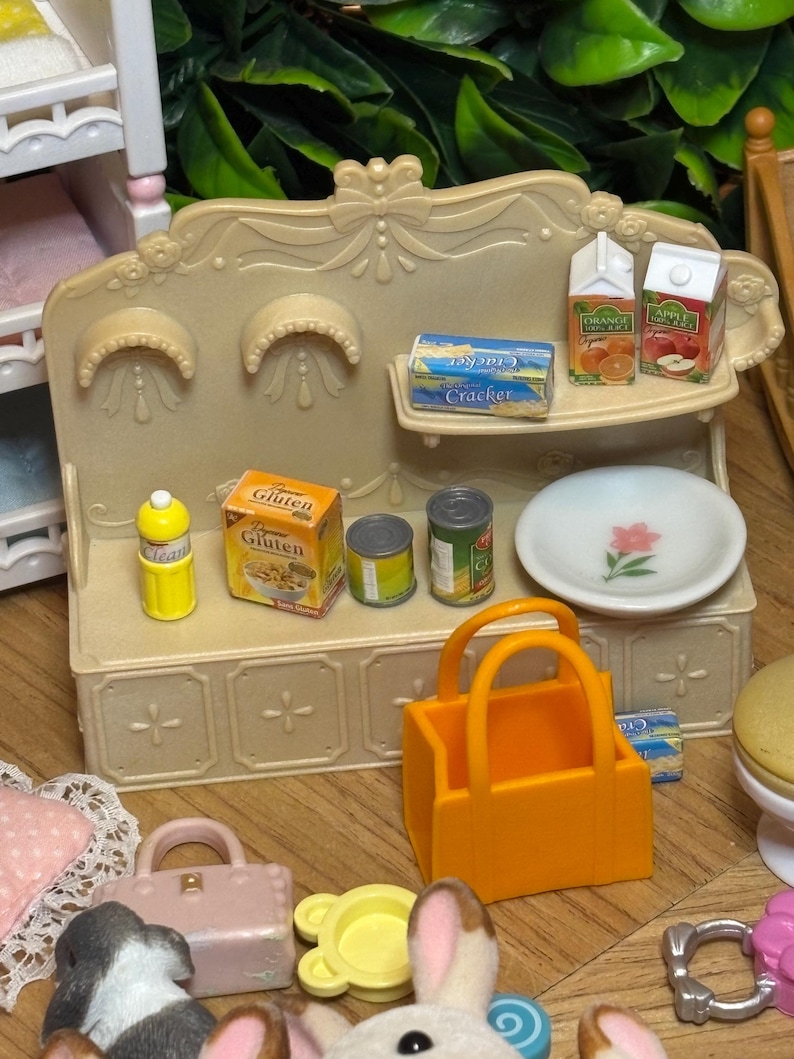 May include: A miniature beige kitchen hutch with a shelf holding two juice cartons and a cracker box. The counter features a yellow bottle, a gluten-free cereal box, two small cans, a white plate with a pink flower, and an orange shopping bag.