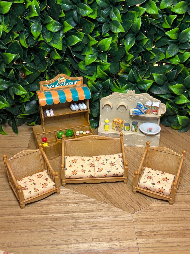 May include: A miniature toy set featuring a brown Food Corner shop stand with an orange and teal striped awning, a beige kitchen unit with various food accessories, and three brown chairs with cream-colored cushions featuring a small floral pattern.