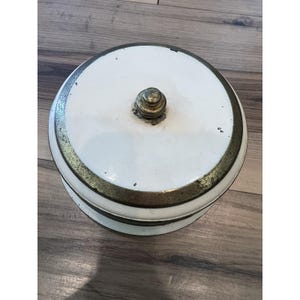 May include: A white, round, vintage container with a brass-colored rim and a brass knob on top. The container has a slightly distressed appearance, suggesting age and use. The design is simple and classic.