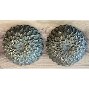 Puede incluir: Dos piezas decorativas de arte mural de metal. Cada pieza es un diseño circular que se asemeja a una flor, con detalles de pétalos en capas. El acabado tiene una pátina de bronce y verde, agregando profundidad y textura. Las piezas se exhiben sobre una superficie de madera.