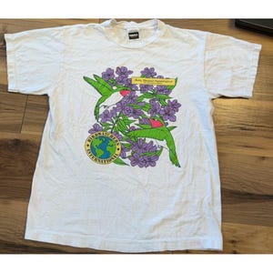 May include: A white short-sleeve t-shirt featuring a graphic of two green hummingbirds with red throats among purple flowers. The design includes a yellow rectangular label reading Ruby-Throated Hummingbird and a circular logo reading Birdwatchers International with a globe illustration.