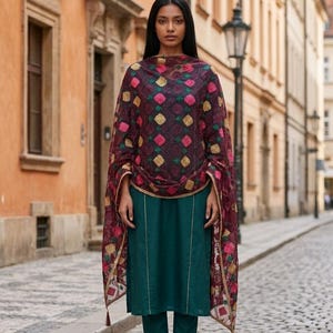 May include: A woman in a teal green salwar kameez suit with a patterned shawl. The shawl has a burgundy background with pink, gold, and green floral designs. The outfit is paired with gold sandals, and the background is a cobbled street.