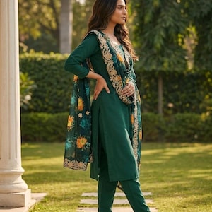 May include: A dark green salwar kameez ensemble. The outfit includes a long-sleeved tunic, matching pants, and a floral-patterned scarf. The scarf features a gold embroidered border and colorful floral designs. The outfit is worn with sandals.
