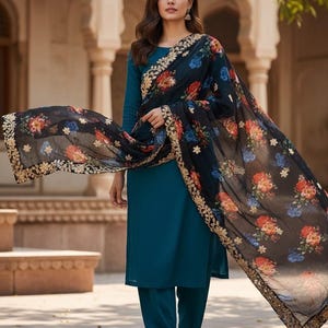 May include: Teal-colored salwar kameez ensemble with a long-sleeved top and matching pants. A black floral scarf with gold trim drapes over the shoulders. The scarf features red, blue, and orange flowers.