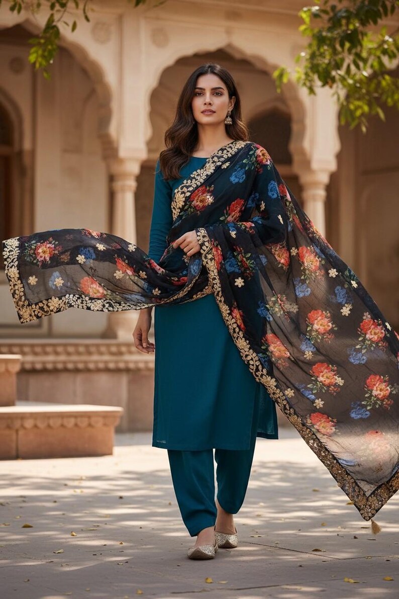 May include: Teal-colored salwar kameez ensemble with a long-sleeved top and matching pants. A black floral scarf with gold trim drapes over the shoulders. The scarf features red, blue, and orange flowers.
