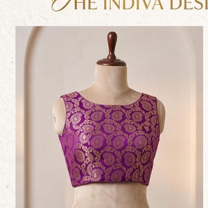 May include: A sleeveless blouse in a vibrant purple hue featuring a repeating gold paisley pattern throughout the fabric. The garment has a wide, rounded neckline and a cropped length, displayed on a mannequin. The text THE INDIVA DESI is visible.