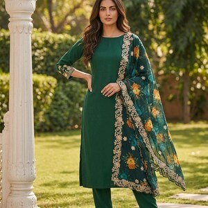 May include: A dark green salwar kameez suit with a matching dupatta. The suit features a long-sleeved tunic and straight-leg pants. The dupatta has a floral print with gold trim. The outfit is worn with gold-colored flats.