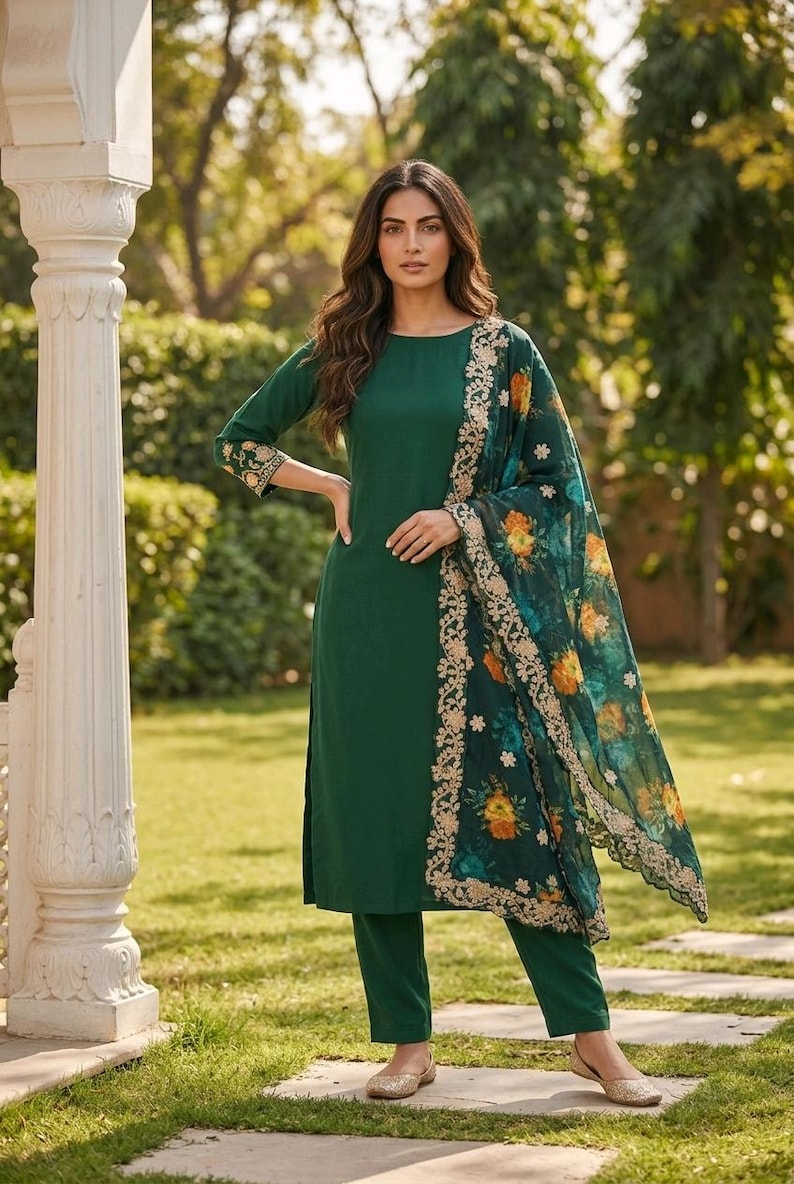May include: A dark green salwar kameez suit with a matching dupatta. The suit features a long-sleeved tunic and straight-leg pants. The dupatta has a floral print with gold trim. The outfit is worn with gold-colored flats.
