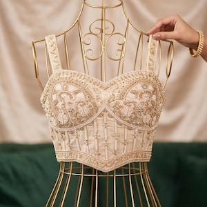 Puede incluir: Un top tipo corsé de color beige con intrincados bordados dorados y cuentas blancas. La prenda presenta un escote corazón, tirantes anchos y un dobladillo en punta, con adornos detallados que cubren toda la superficie frontal.